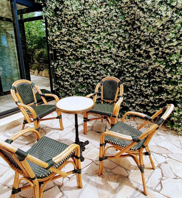 A set of wicker chairs and a table arranged on a tiled patio with a backdrop of dense flowering bushes.