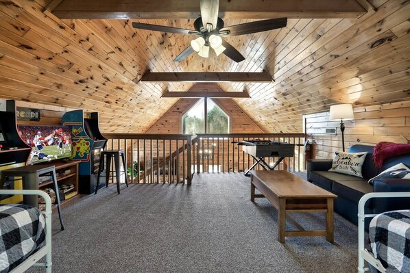 A cozy attic room with a wooden ceiling, a ceiling fan, a foosball table, a sofa, and a TV displaying a sports game. - Award winner| Dollywood |Arcades|Hot tub |GSMNP (Sevierville)
