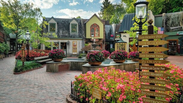 A quaint village square with cobblestone paths, flower beds, and a signpost with various business names. - Award winner| Dollywood |Arcades|Hot tub |GSMNP (Sevierville)