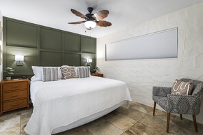 Ein Schlafzimmer mit einem großen Bett, einem Deckenventilator, einem Nachttisch mit Lampe, einem Sessel und einem Fenster mit Jalousien.