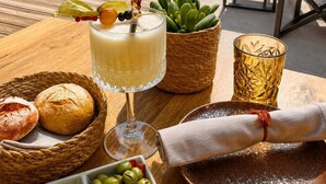 A beachside dining setup with a table, chairs, a glass of drink, bread, olives, and a cactus plant.