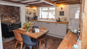 A kitchen with wooden beams, a brick wall, and a window. There is a table with chairs, a stove, and various kitchen items.