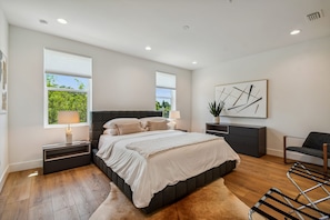 Une chambre à coucher moderne avec un grand lit, des tables de chevet, une commode, une chaise et une grande fenêtre avec des stores.