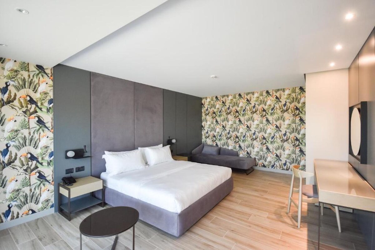 A modern hotel room with a large bed, a desk, and a chair. There is a small table and a nightstand with a lamp. The walls have a tropical wallpaper design.