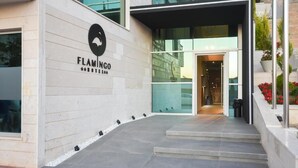 Ein moderner Gebäudeeingang mit Glastür, einem Schild mit der Aufschrift „FLAMINGO“ und einer kleinen Außenanlage mit Stufen und einem Blumenbeet mit roten Blüten.