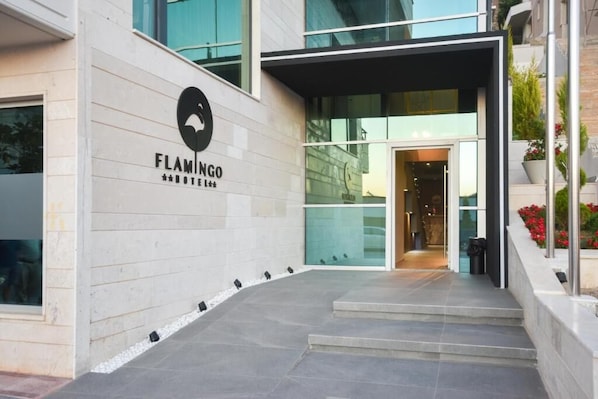 A modern building entrance with a glass door, a sign reading "FLAMINGO," and a small outdoor area with steps and a planter with red flowers.