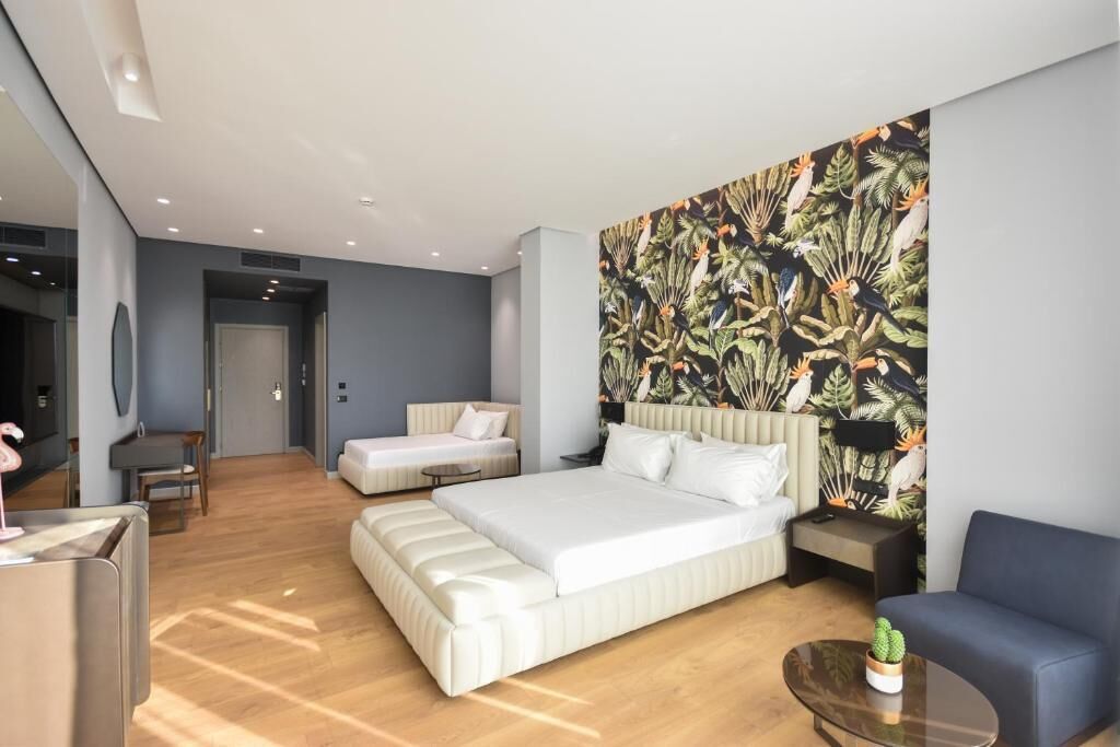A modern hotel room with a large bed, a sofa, a small table, and a wall with a tropical leaf pattern.