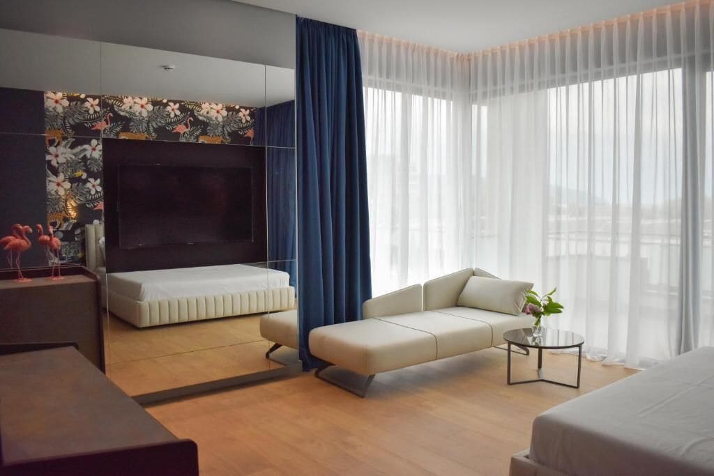 A modern hotel room with a large flat-screen TV, a sofa, a bed, and a small table with a vase.
