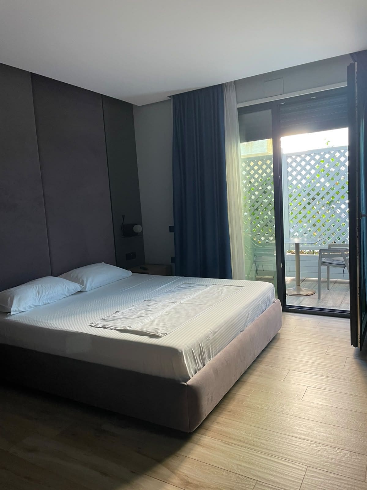 A bedroom with a bed, a nightstand, a window with curtains, and a view of an outdoor area.