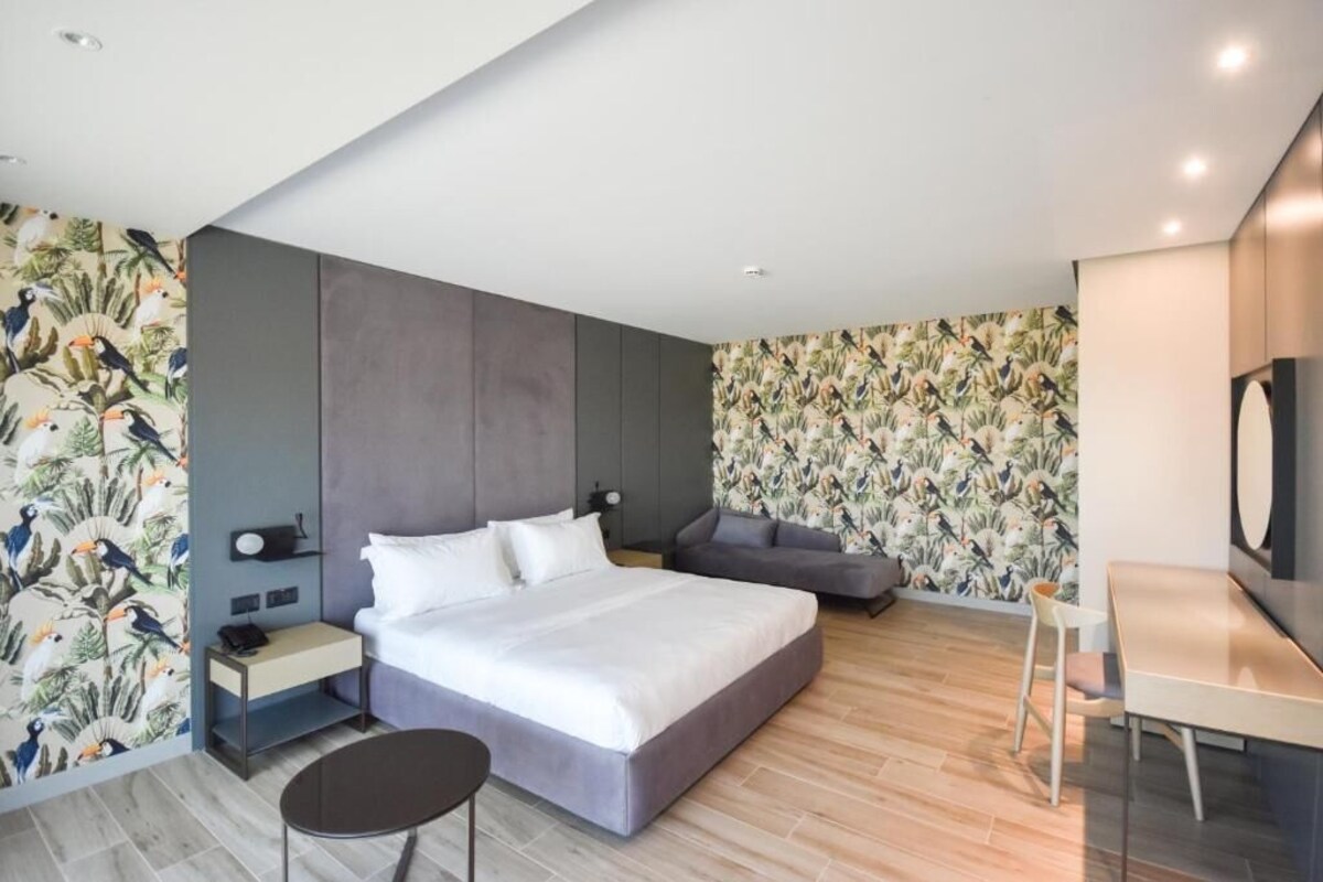 A modern hotel room with a large bed, a desk, and a chair. There is a small table and a nightstand with a lamp.