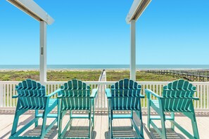A wooden deck with four green Adirondack chairs facing a beach.