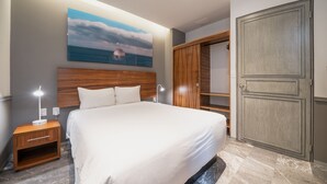 A neatly made bed with a wooden headboard, a bedside table with a lamp, a large painting of a seascape, and a wardrobe with a double door. - Condo Jade - Singular Joy Residences (Playa del Carmen)