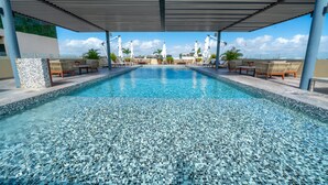 A swimming pool with tiled edges, surrounded by lounge chairs and umbrellas, under a covered area with a view of the sky. - Condo Jade - Singular Joy Residences (Playa del Carmen)