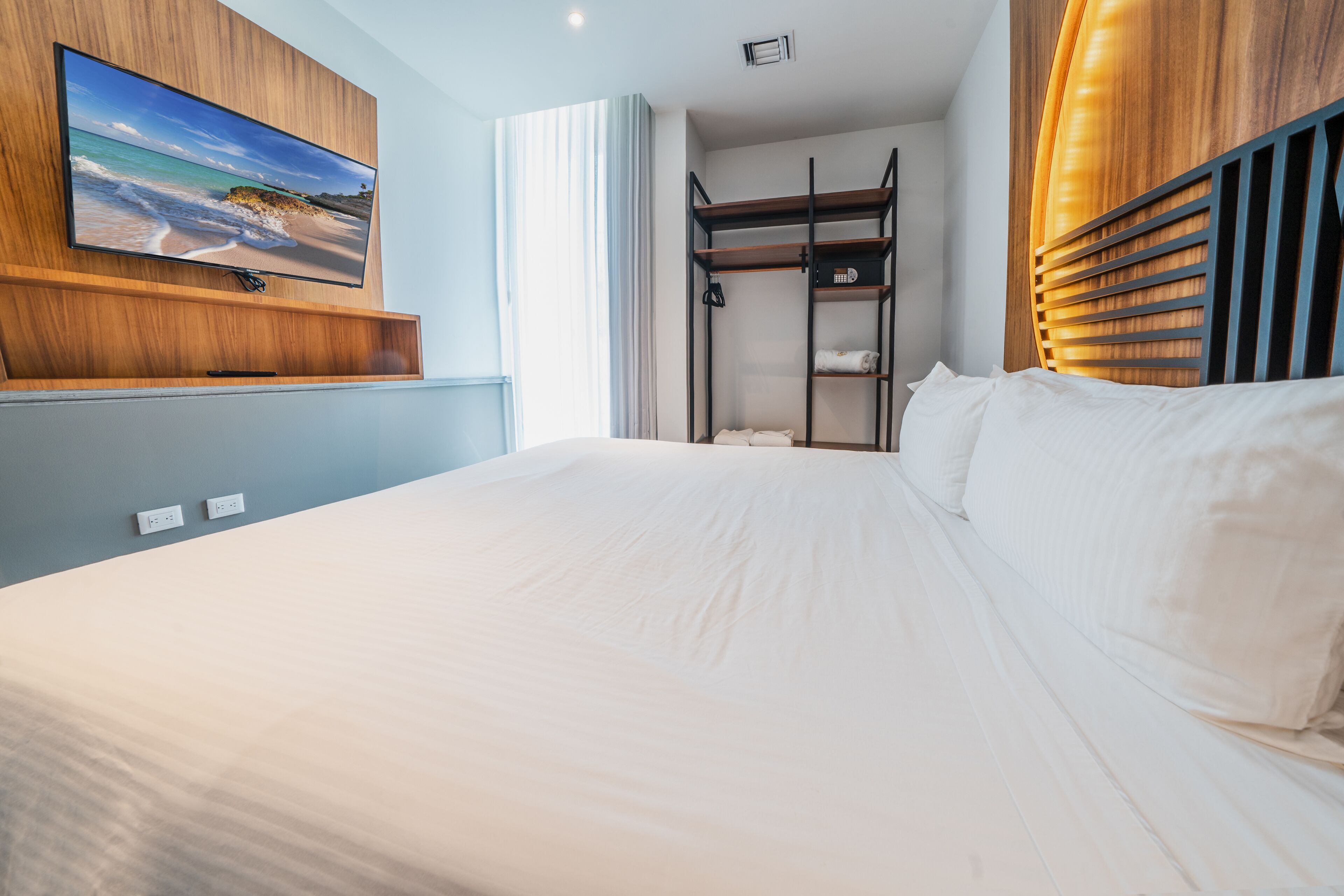 A modern hotel room with a large bed, a mounted TV, a built-in shelf, and a window with curtains.
