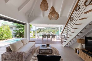 A modern living room with a sofa, coffee table, and TV, overlooking a pool and outdoor dining area. - Modern villa, a 3-minute walk from St. Jean Beach, shops, and restaurants. (SAINT-JEAN)