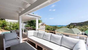 Ein überdachter Außenbereich mit Pool, heller Möbel und Blick auf Meer und Hügel.