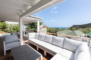A covered outdoor seating area with a pool, white furniture, and a view of the sea and hills.