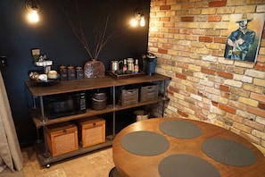 Una cocina con pared de ladrillo, una mesa de madera con individuales, y estantes con utensilios de cocina y especias.