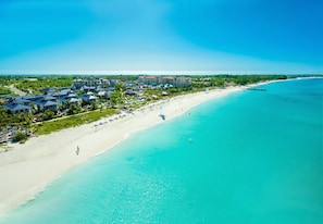 Vue aérienne d’une station balnéaire avec des propriétés situées en bord de mer, une plage de sable et des eaux turquoise cristallines.