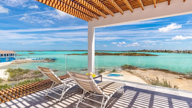 Un balcon avec deux chaises longues, une table avec une boisson, et une vue sur des eaux turquoise et des îles lointaines.