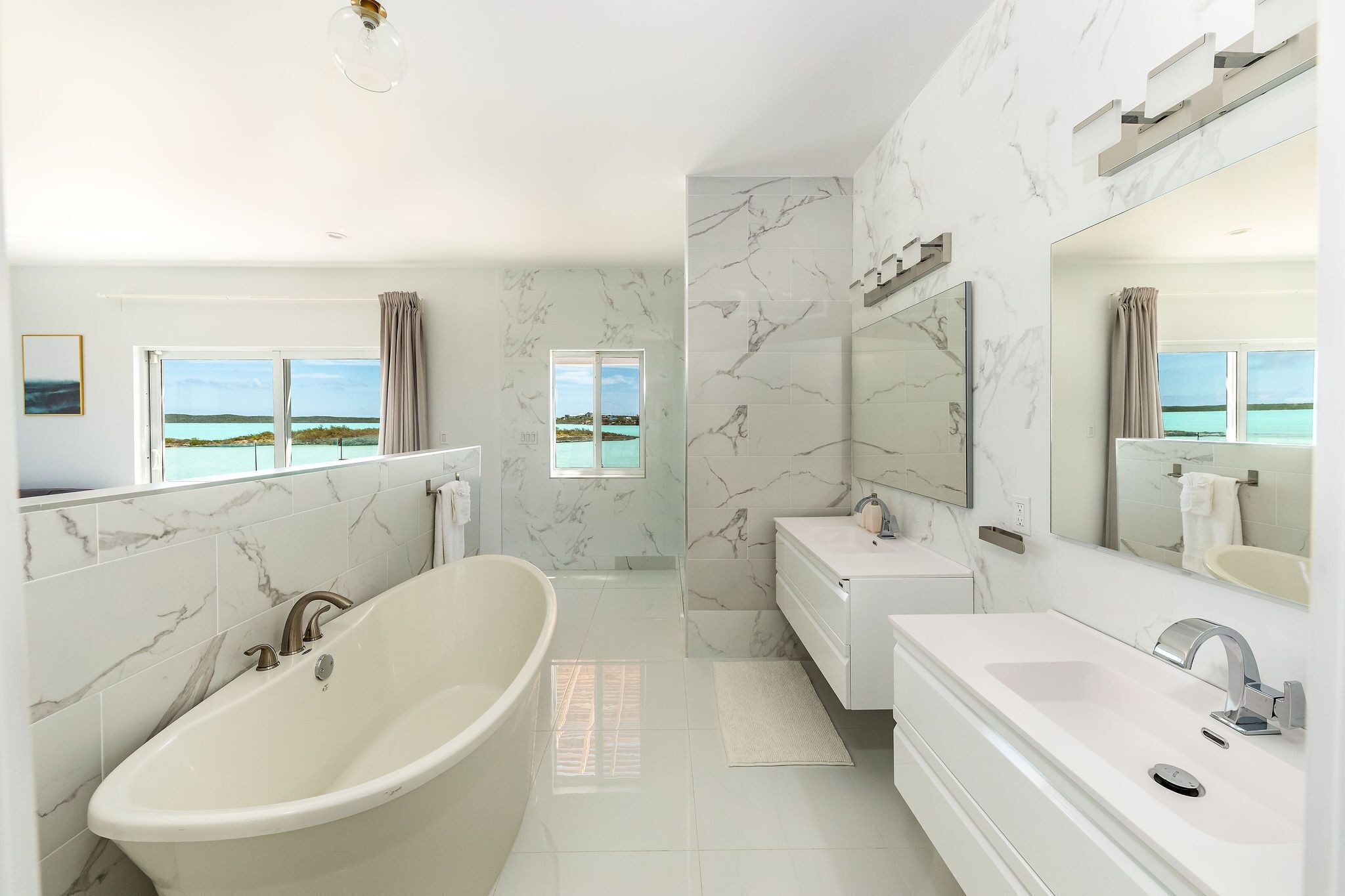 Ein modernes Badezimmer mit einer Badewanne, einem Waschtisch und einem großen Spiegel. Fenster bieten Blick aufs Meer.