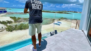 Un homme se tient près d’une piscine, avec vue sur une plage et des eaux turquoise.