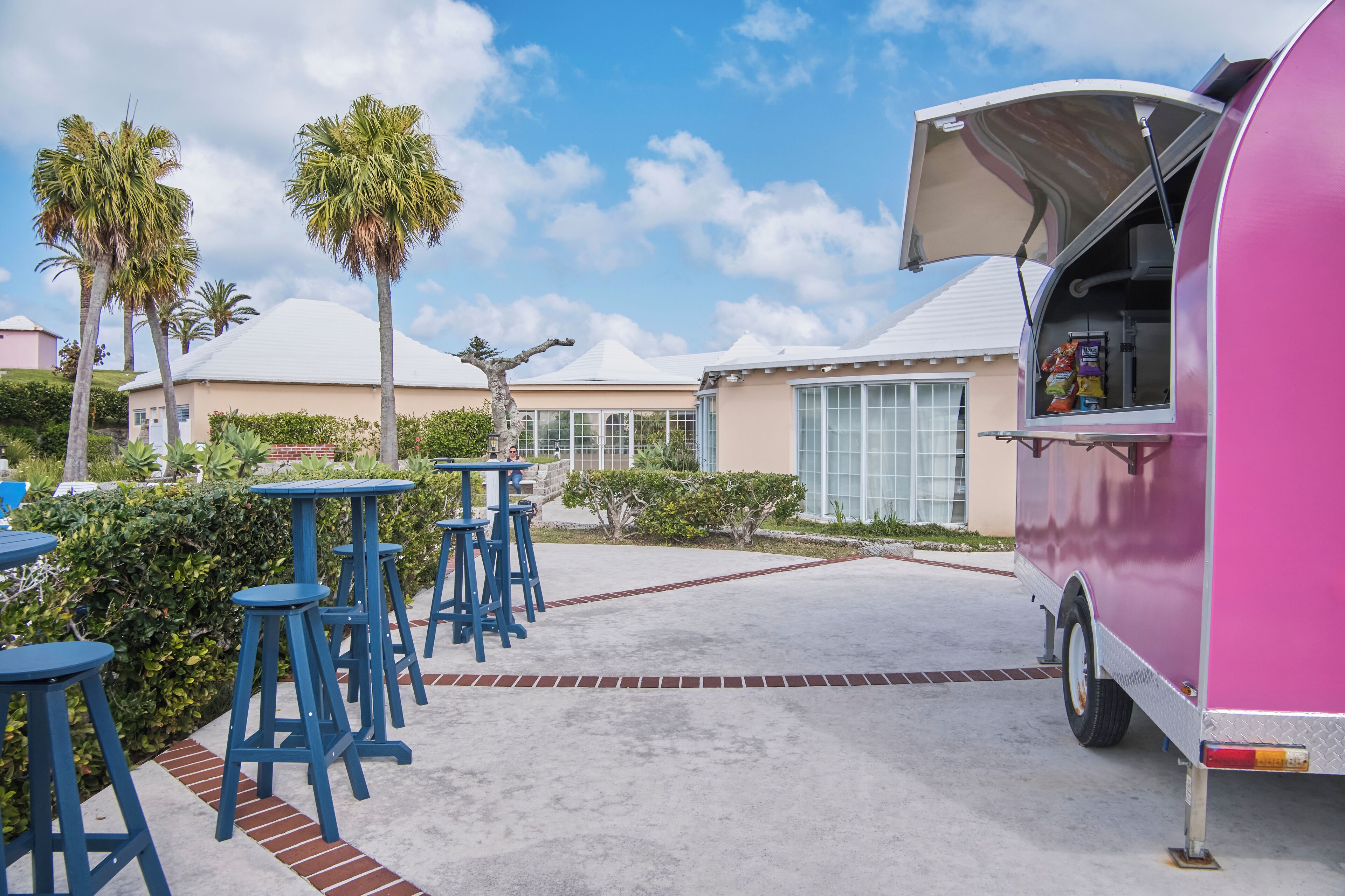 Un food truck rosa amb la tenda estesa, estacionat en una zona asfaltada amb tamborets blaus i palmeres al fons.