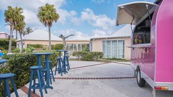 A pink food truck with its awning open, parked in a paved area with blue bar stools and palm trees in the background.