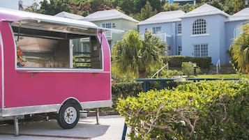 Un camión de comida rosa estacionado frente a una hilera de casas azules con un jardín cuidado y un cielo despejado.