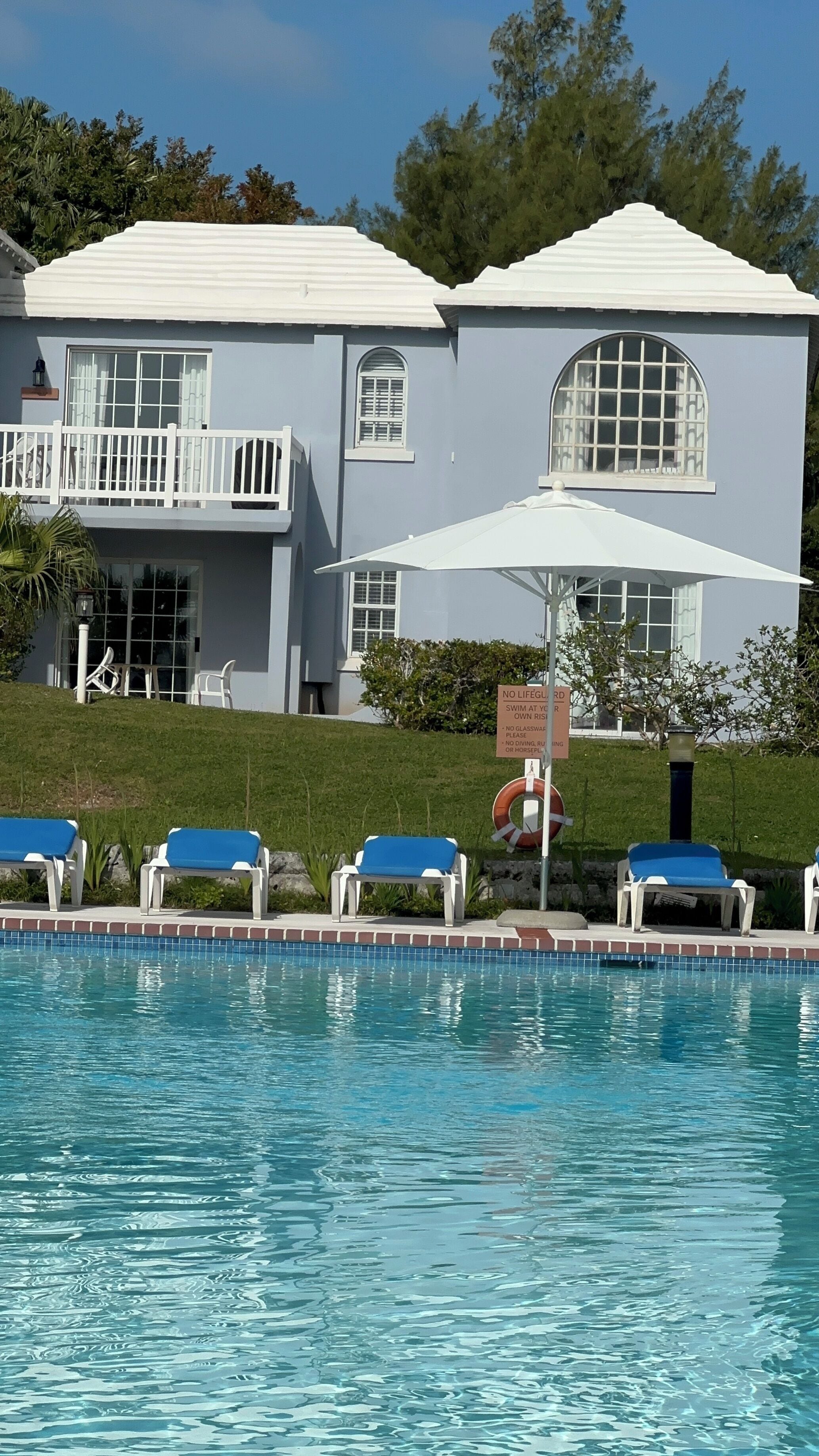 A pool area with lounge chairs, a lifeguard sign, and a building with large windows and a balcony.