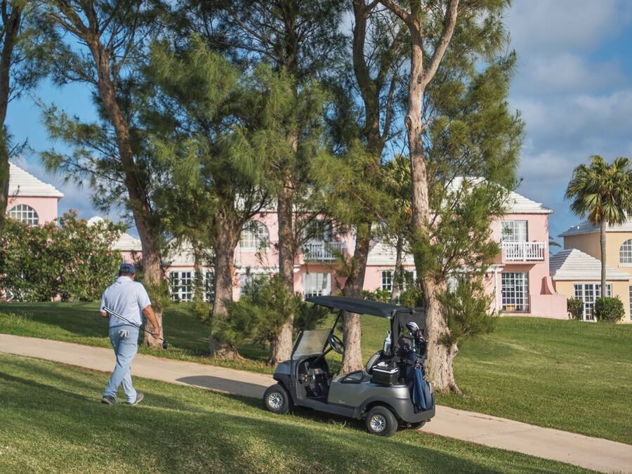 A golf cart on a path with a man walking behind it. There are trees and buildings in the background.