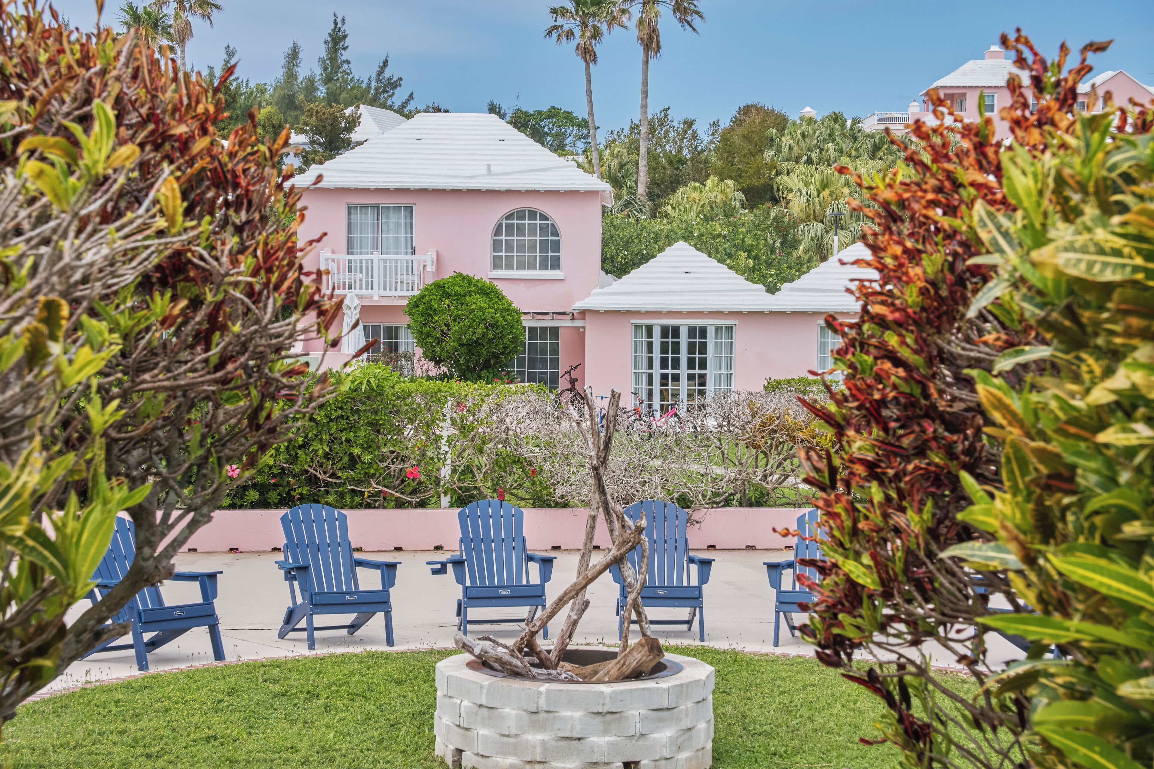 Una casa rosa con techo blanco, rodeada de sillas Adirondack azules y un árbol en una jardinera.