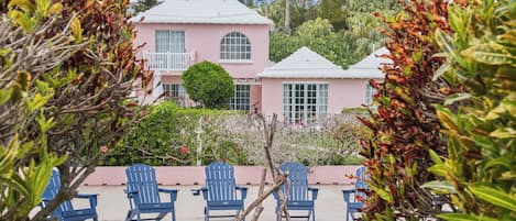 Una casa rosa con techo blanco, rodeada de sillas Adirondack azules y un árbol en una jardinera.