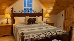 Ein Schlafzimmer mit schrägverdeckter Decke, einem Bettgestell aus Holz, zwei Nachttischlampen, einem Fenster und einer schwarzen Lederbank.