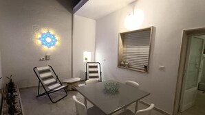 A small, well-lit room with a glass-top table, white chairs, a folding chair, a wall-mounted light fixture, and a window with blinds. - Javec patio 2 minutes from town center, 10 minutes from the sea and (Carovigno)