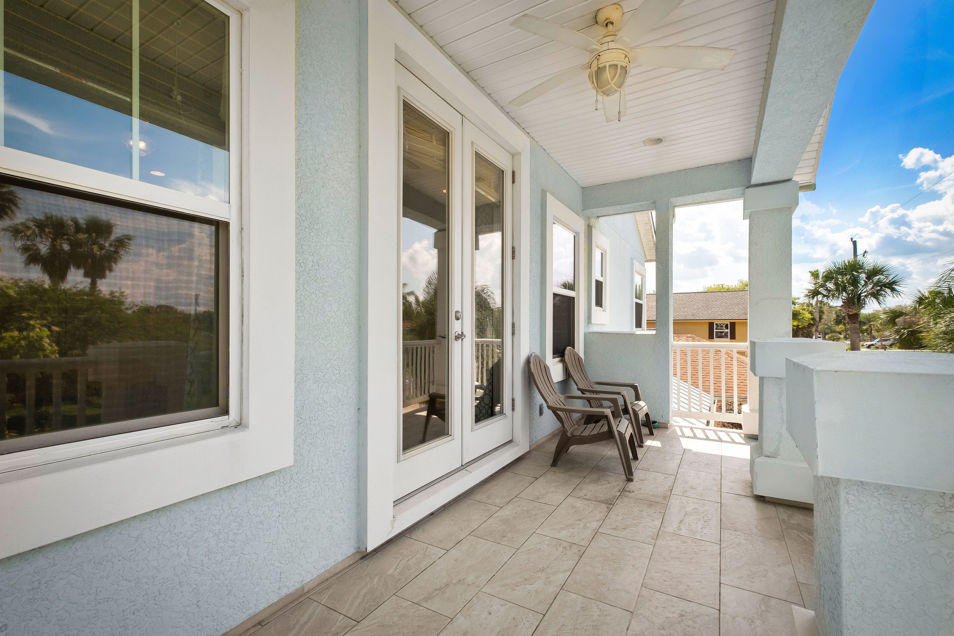 A covered patio with white walls and a ceiling fan, featuring a seating area with chairs and a view of the surrounding neighborhood.
