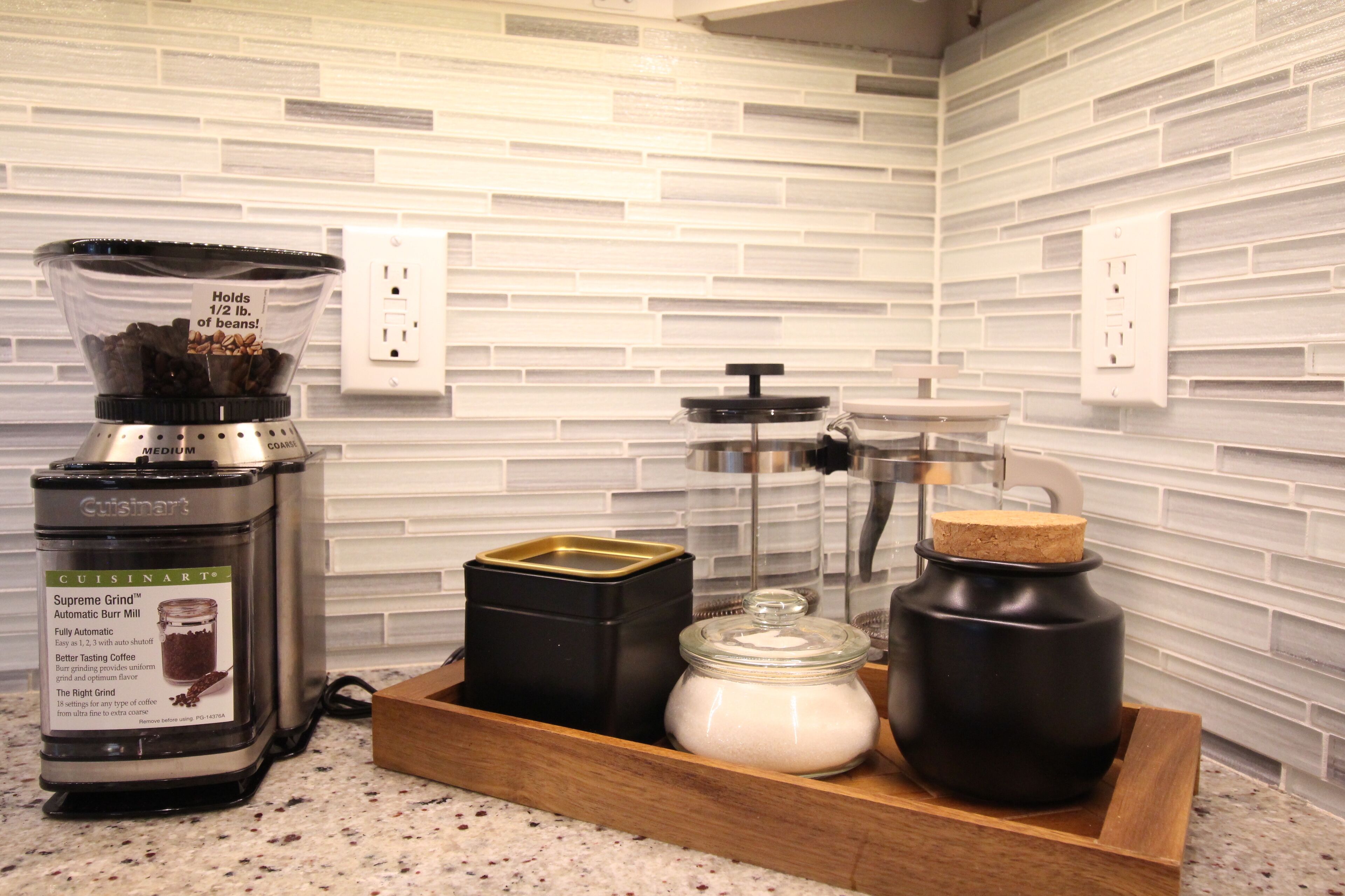 Une cuisine avec un moulin à café, un pot de sucre et un récipient noir posé sur un plateau en bois.