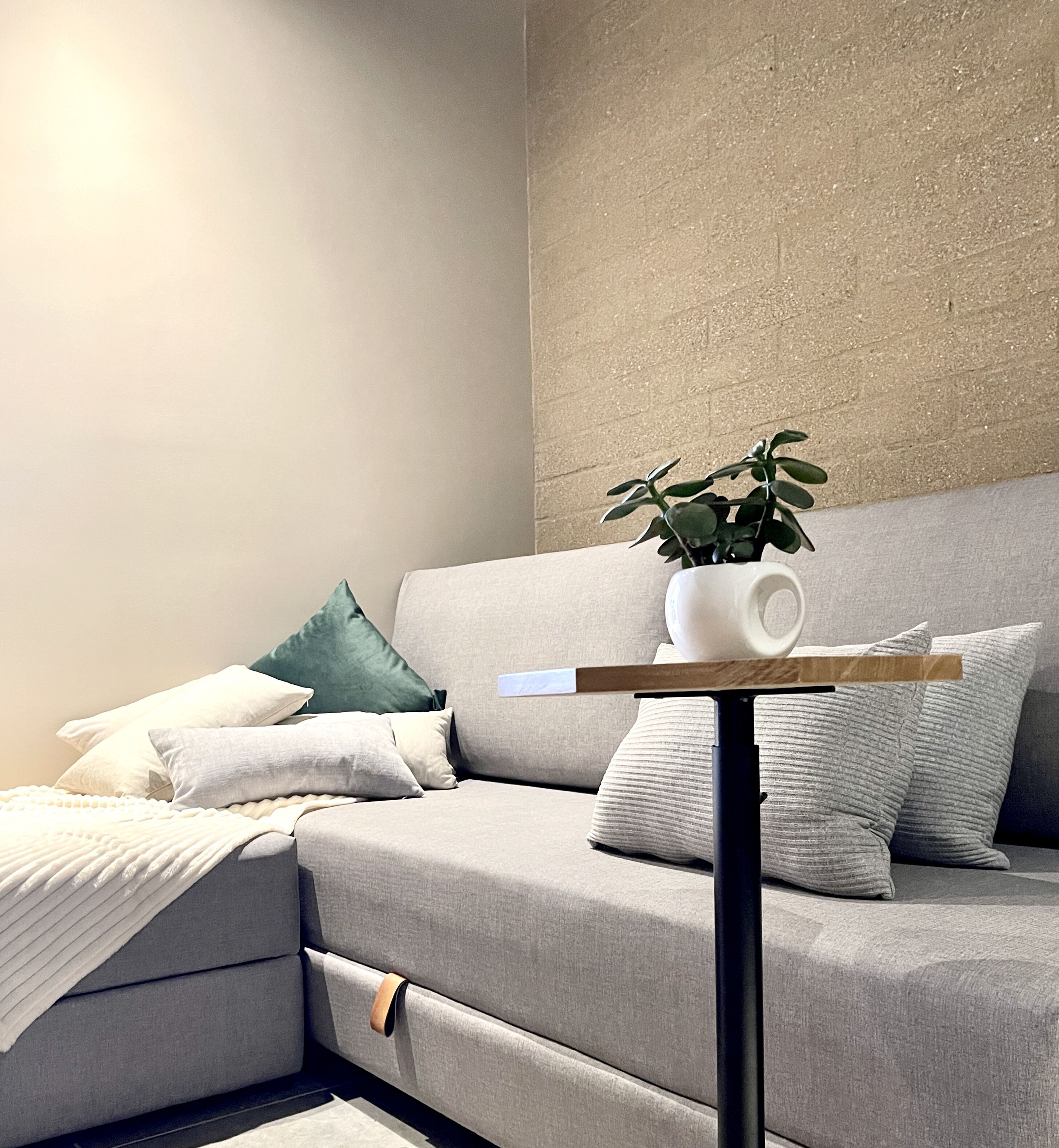 Una sala de estar moderna con un sofá gris, almohadas verdes y blancas, un jarrón blanco con una planta y una mesita de madera con un objeto blanco encima.