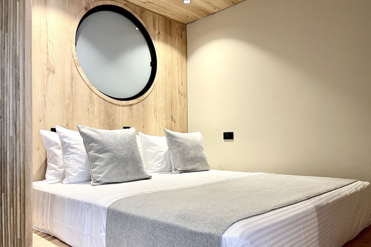 A bedroom with a wooden headboard, a round mirror, and a bed with white bedding and gray throw.