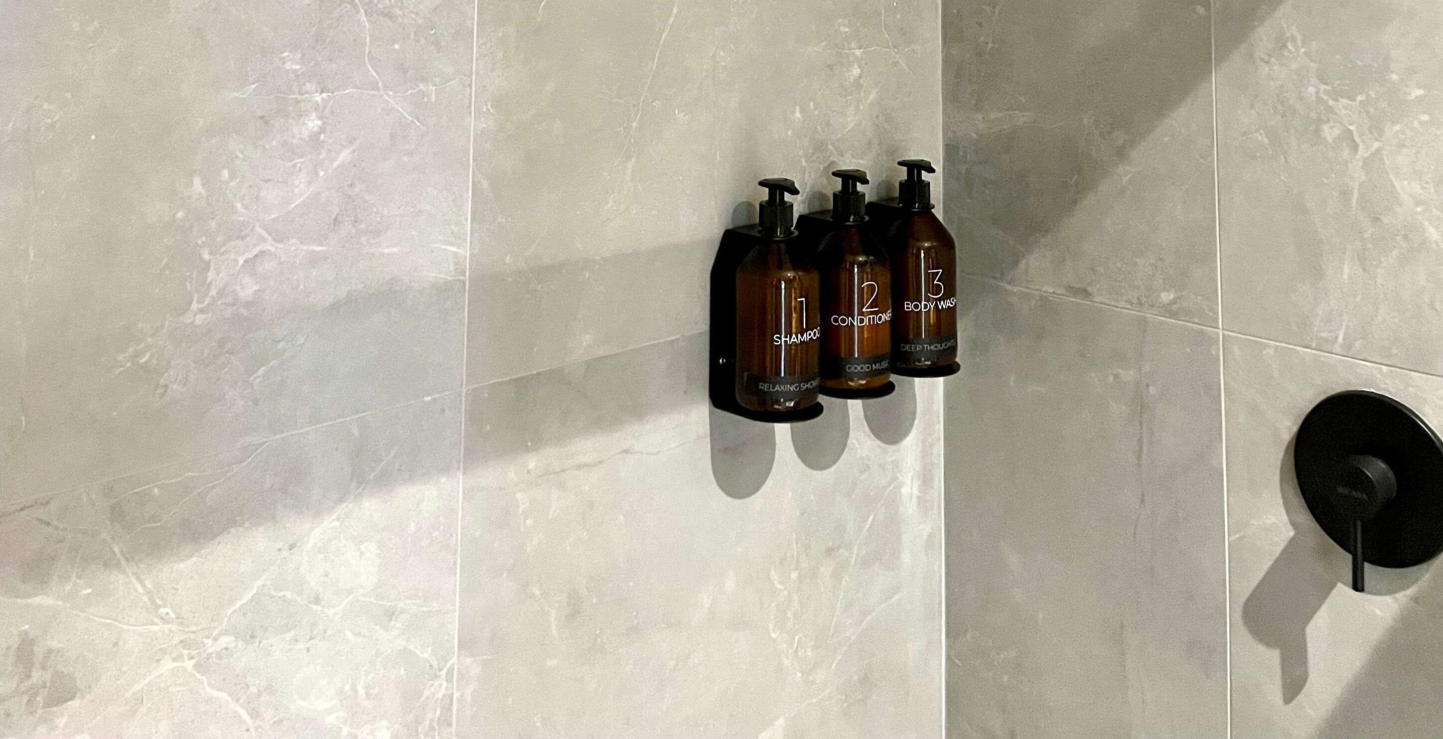 A bathroom with three bottles of body wash and shampoo on a tiled wall.