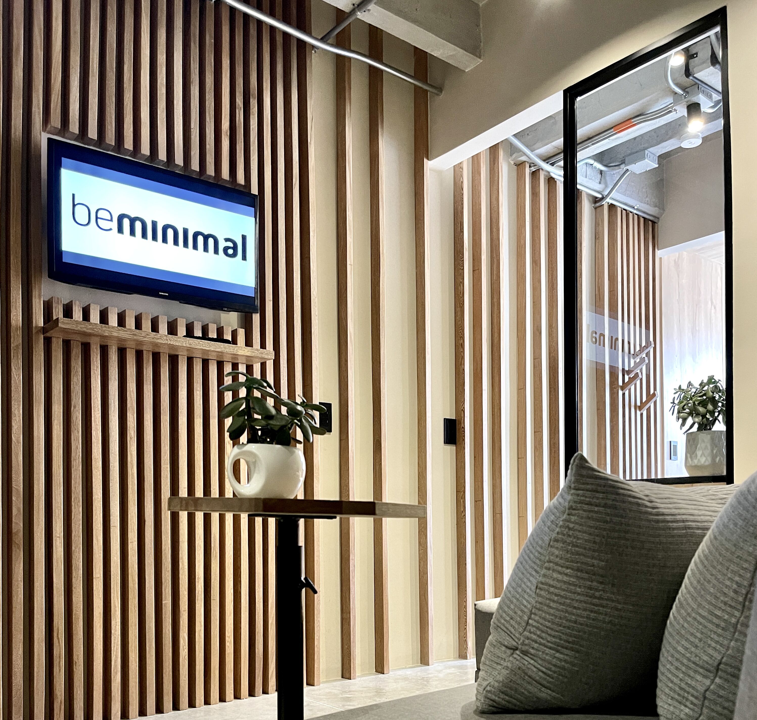 A modern interior with a television displaying 'bemminimal', a wooden slatted wall, a small shelf with a plant, and a couch with cushions.