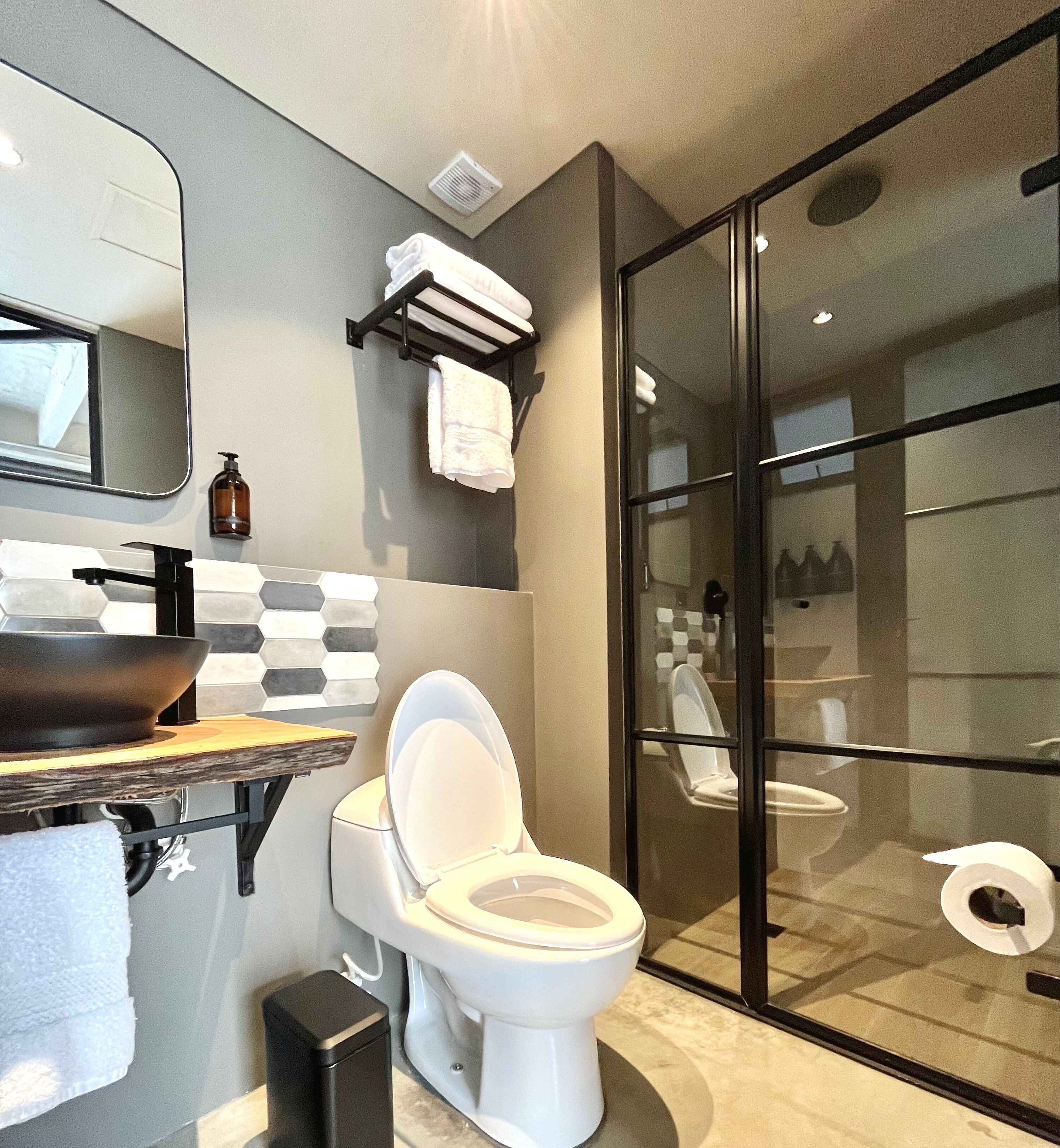 A modern bathroom with a large mirror, a black sink, a toilet, and a glass-enclosed shower area.