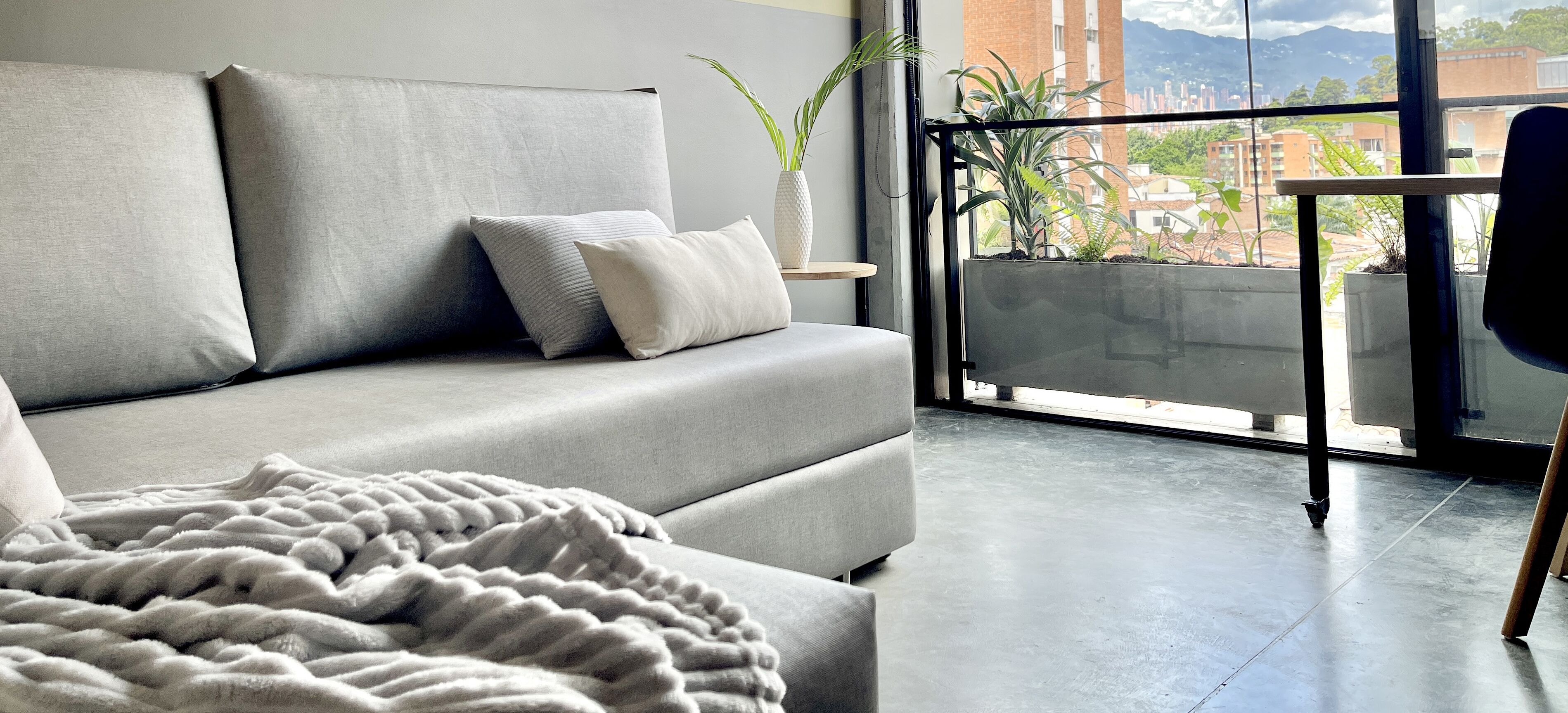 Un salon moderne avec un canapé gris, un plaid blanc et une vue sur un balcon agrémenté de plantes et de bâtiments.