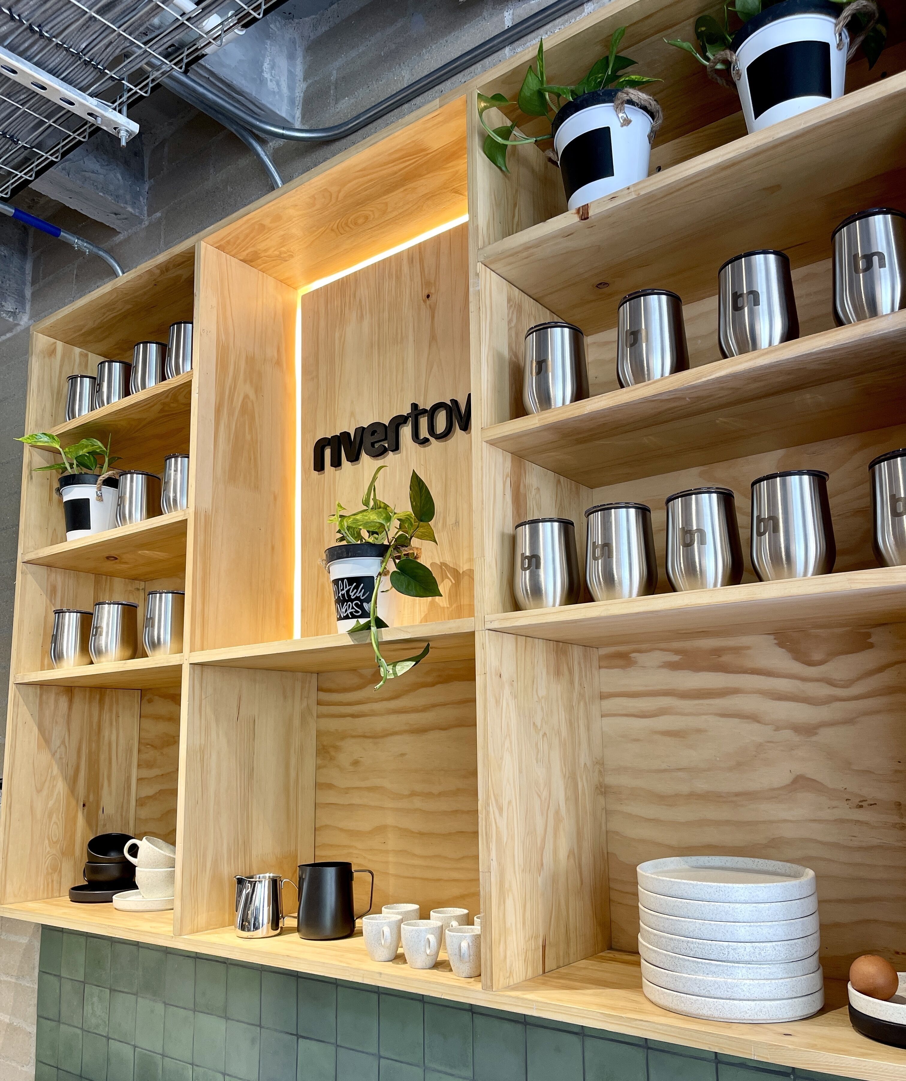 Una estantería de madera con varios compartimentos, algunos con plantas en macetas y otros con tazas de acero inoxidable.