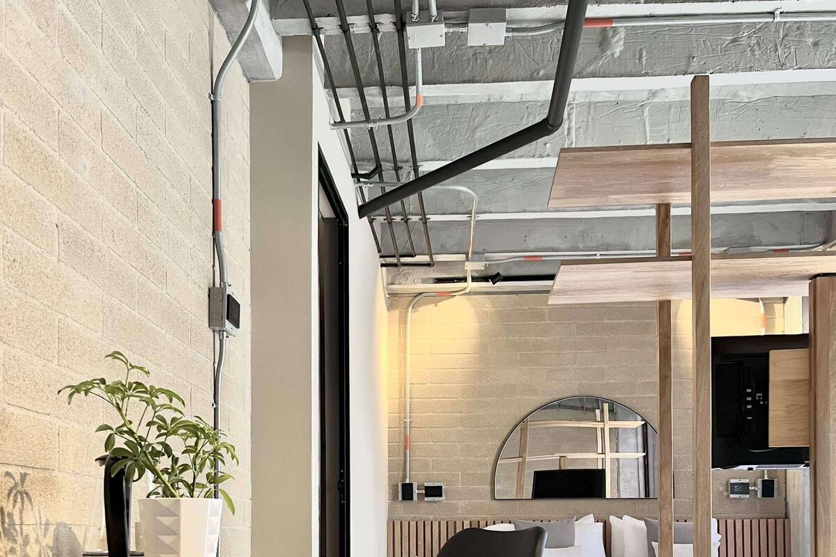 A modern, open-plan room with a wooden desk, a black chair, a bed with a wooden headboard, and a kitchen area in the background.