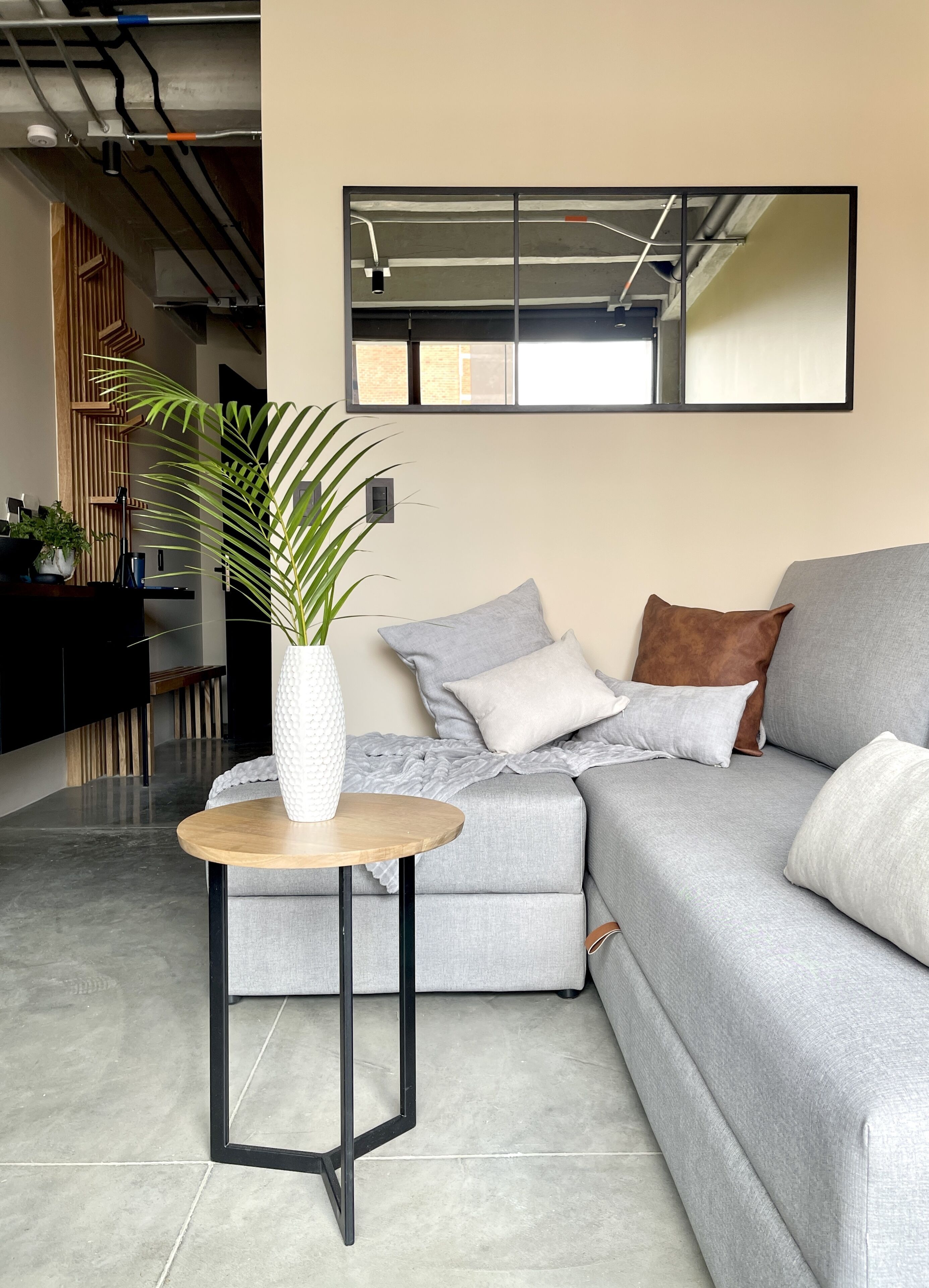 A modern living room with a grey sofa, a round wooden table with a white vase, and a large mirror on the wall.
