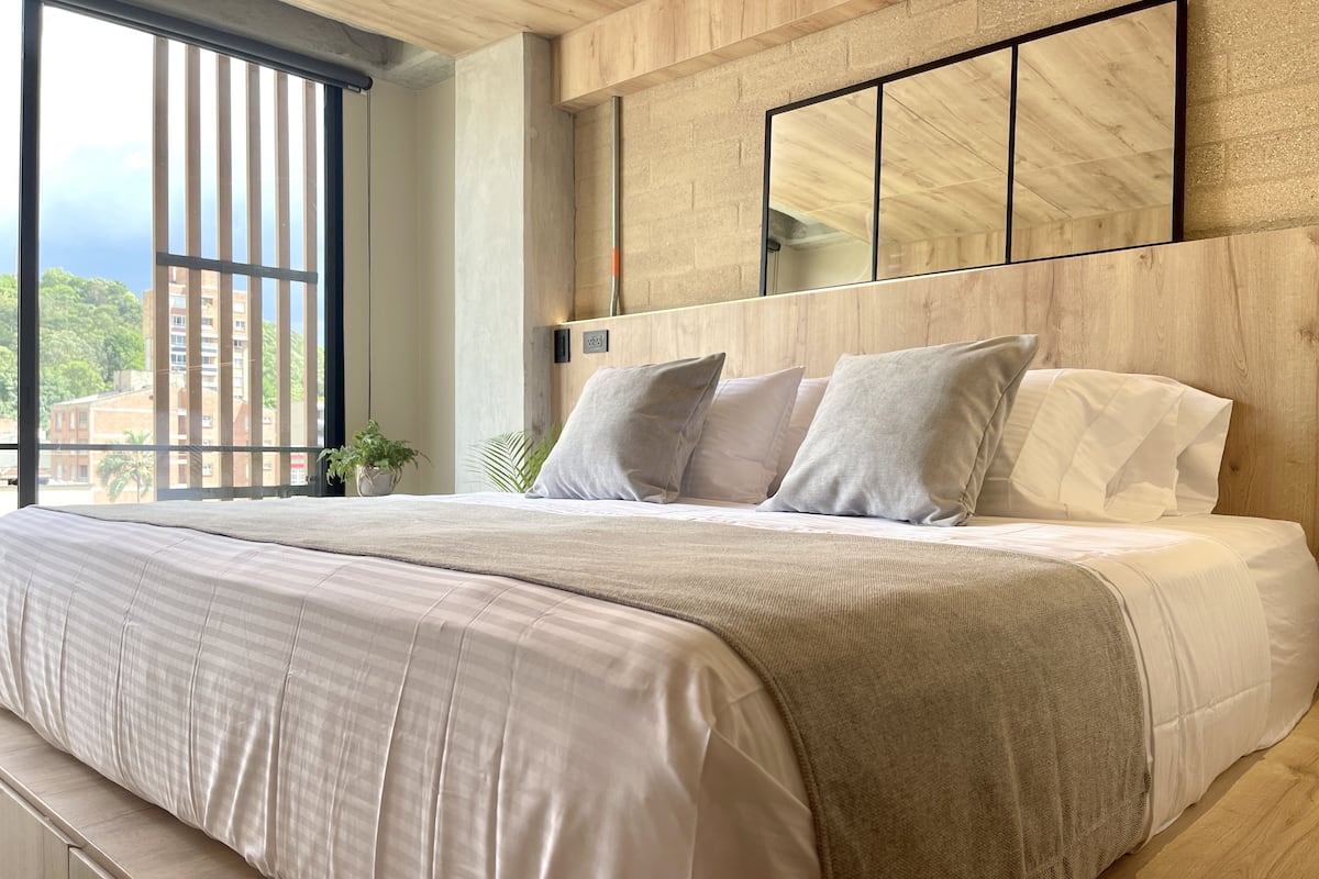 A modern bedroom with a large bed, a wooden headboard, and a view of the city through large windows.