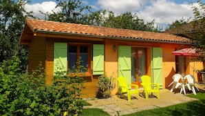 A small, single-story wooden house with a tiled roof, green shutters, and a patio with two chairs. - Gite in the mountains (Seix)