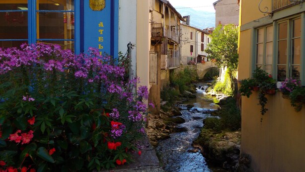 A narrow street with a building labeled "MIXTE" on the left, a stone bridge, and a stream running through the center. - Gite in the mountains (Seix)