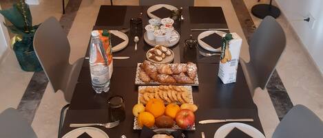 Una mesa de comedor dispuesta con diversos alimentos y utensilios, rodeada de sillas, en una habitación con pared de piedra y una cama al fondo.